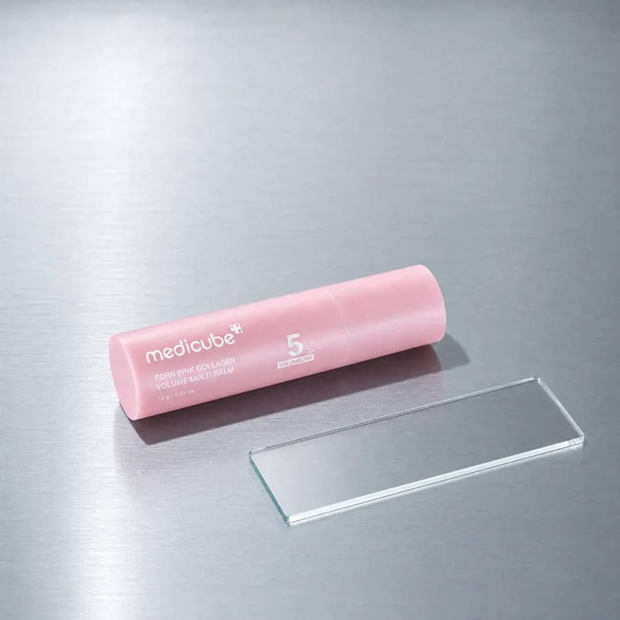 The medicube balm stick lying on a brushed steel surface beside a clear glass slide in cool laboratory light