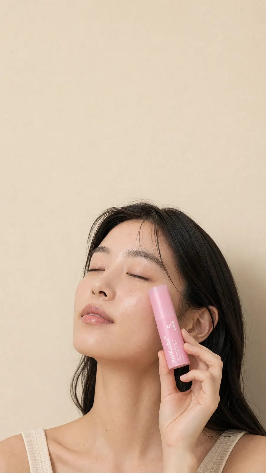 A woman pressing the pink medicube PDRN Pink Collagen Volume Multi Balm stick to her cheekbone beside a bare cream plaster wall